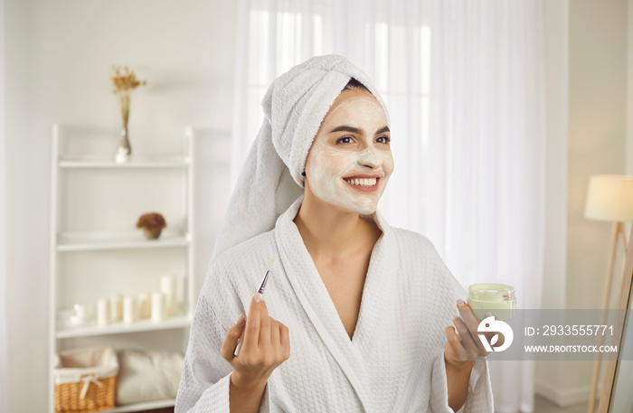 Women’s cosmetic care. Portrait of beautiful young woman applying cosmetic face mask during morning beauty routine. Smiling woman in white bathrobe and towel holding container with mask and brush.