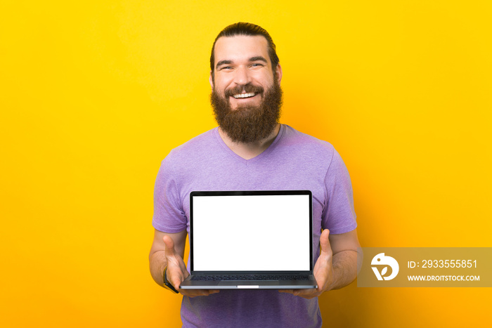 Wonderful bearded man is holding a blank screen laptop to the camera over yellow background.