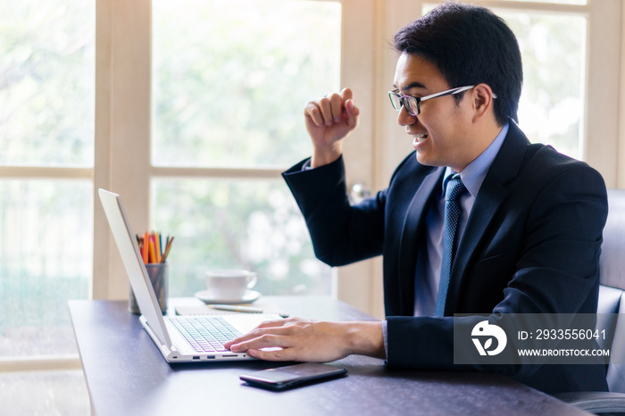 Young happy businessman using his laptop and glad while Raise a fist up on office desk. Portrait of business executive man using laptop in office with successful.