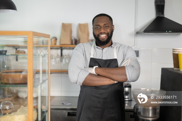 Cheerful and friendly African-American small business owner, cafe manager, bakery staff stands in confident pose with arms crossed and looks at the camera