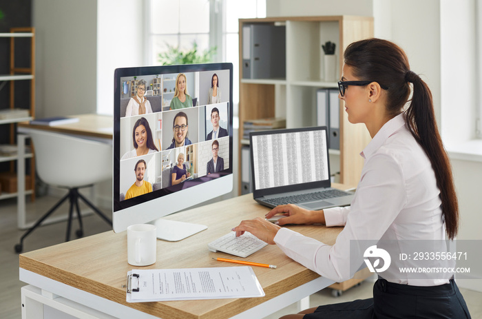 Female web conferencing administrator sitting at office table with desktop computer during virtual team meeting of company workers. Online business communication, telework and remote teamwork concepts