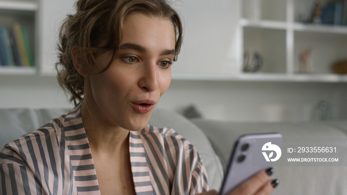 Surprised woman using smartphone reading messages in pajamas at home closeup.