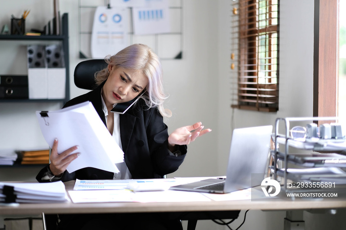 Asian  woman cover her face with hand and feel upset while talk on mobile phone with customer in front of laptop computer on desk at office,Stress office lifestyle concept.