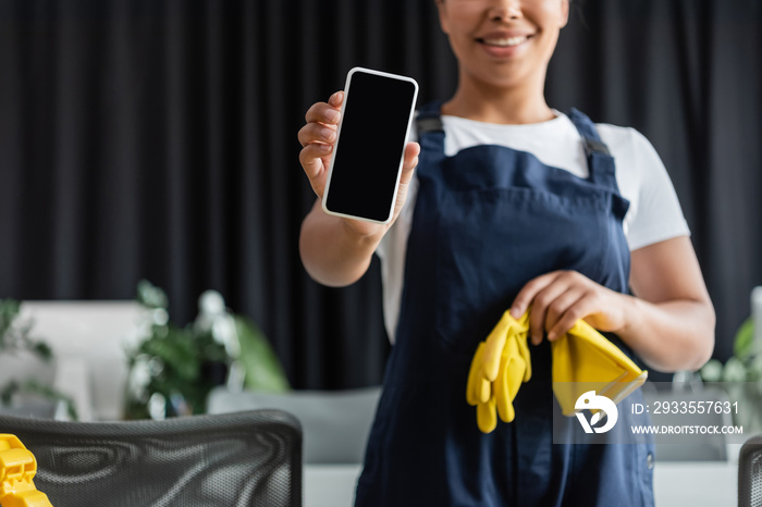 cropped view of smiling mixed race woman holding rubber gloves and smartphone with blank screen.