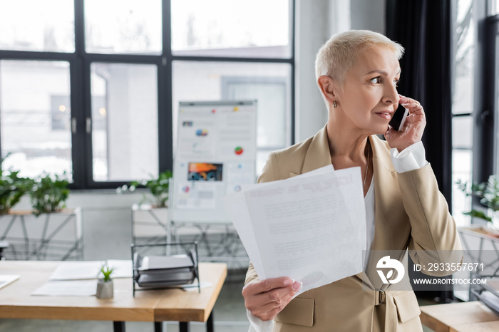 stylish banker with documents looking away while talking on smartphone in blurred office.