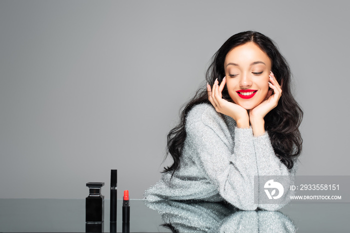 happy woman with red lips looking at bottle with perfume and decorative cosmetics isolated on grey