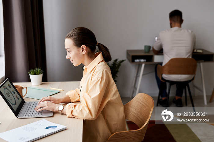 Side view of young confident brunette businesswoman looking at graphic data on laptop screen while sitting by workplace at home