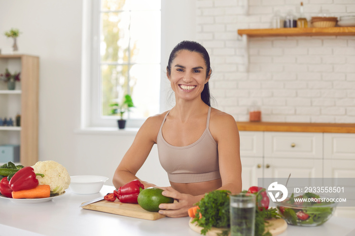 Portrait of happy female cooking healthy vegan food. Fit young woman in sportswear looking at camera and smiling, sitting at kitchen table with cutting board and lots of vegetables for vegetarian meal