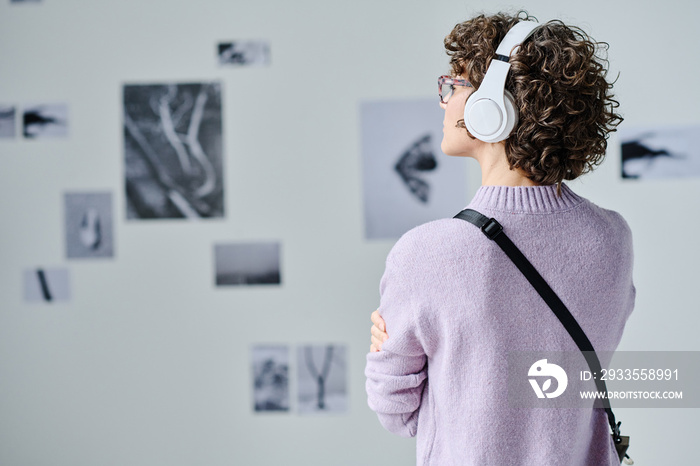 Rear view of young visitor in wireless headphones listening to guide while examining pictures on the wall