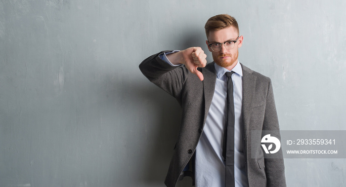 Young redhead elegant business man over grey grunge wall with angry face, negative sign showing dislike with thumbs down, rejection concept