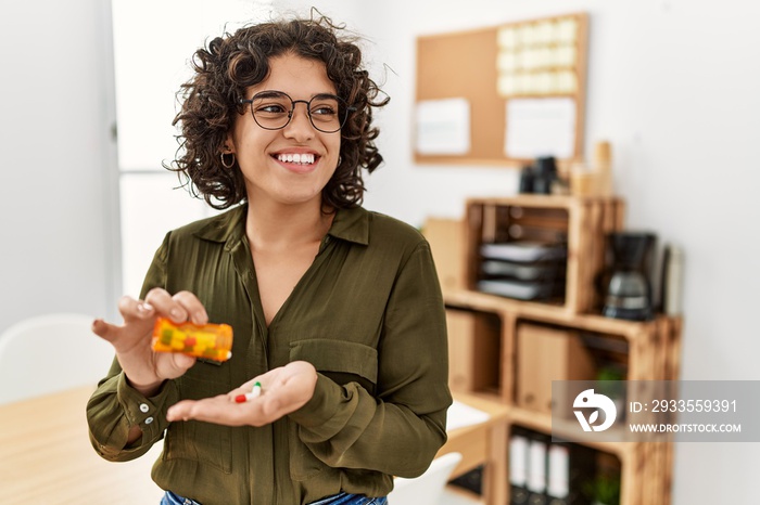 Young hispanic woman smiling confident taking pills at office