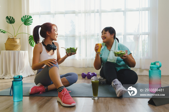 Fitness and Healthy food concept. Two Asian women body size is different in sportswear sitting smiling enjoys while eating healthy food together and happiness after fitness exercising at home.