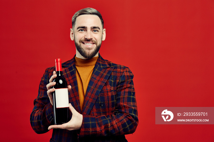 Waist up portrait of elegant bearded man holding bottle of wine and smiling at camera while posing against red background, copy space