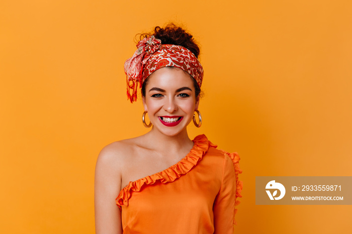 Young girl in great mood is smiling on orange background. Stylish dark-haired lady in orange blouse and scarf on her head looks into camera