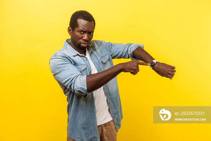 Look at the time! Portrait of irritated impatient man in denim casual shirt pointing at wristwatches, nervous about being late for meeting, deadline. indoor studio shot isolated on yellow background