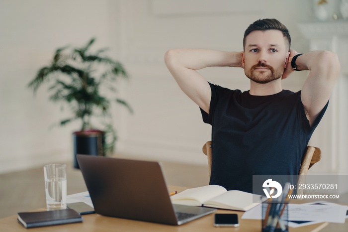 Pensive male freelancer takes break keeps hands behind head poses at desktop works on laptop computer has thoughtful expression thinks about future plans plans week dressed in black t shirt.