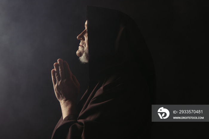 side view of medieval monk in hooded robe praying on black with smoke.