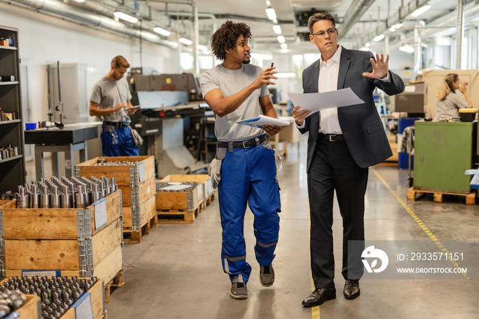 Mid adult engineer and African American worker communicating while walking through factory plant.