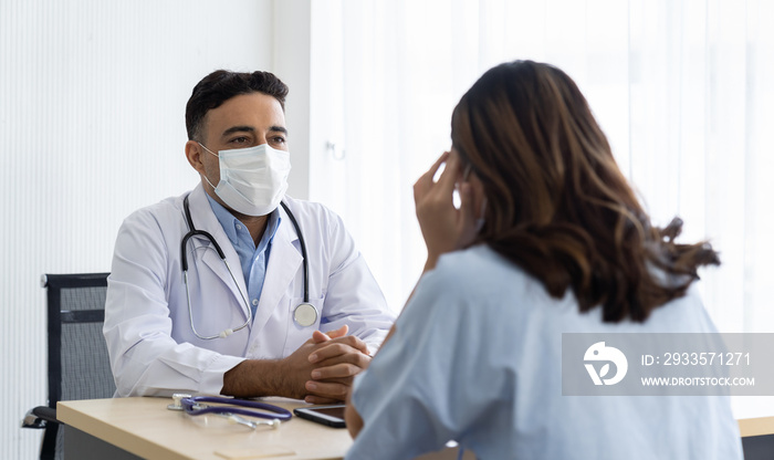 Male doctor wearing face mask discussing with woman patient while sitting on the table in the office at the hospital