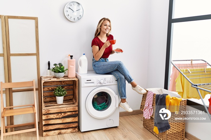 Young caucasian girl drinking coffee waiting for laundry sitting on whasing machine at home.