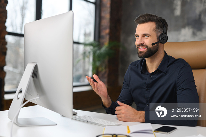 Cheerful guy using handsfree headset and computer to talking online at his workplace. Confident man sitting at the office desk and working with pleasant smile