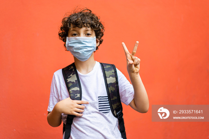 Cute schoolboy with backpack and face mask isolated on a vivid background making V, victory gesture. Back to school in coronavirus time concept.