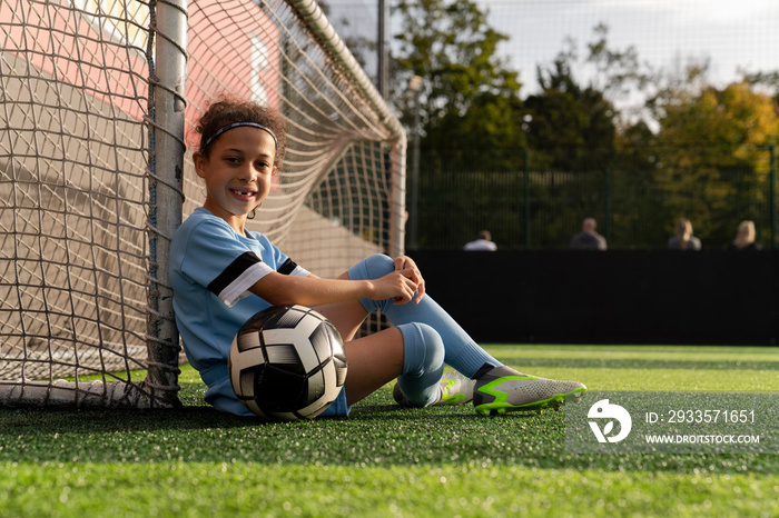 Portrait of girl (6-7) with ball sitting next to soccer goal