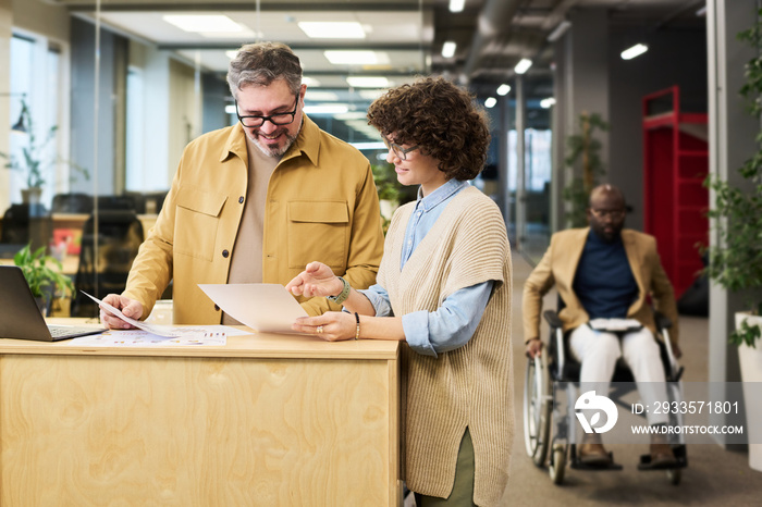 Young female analyst explaining financial data to colleague at meeting while pointing at paper document with diagrams and charts