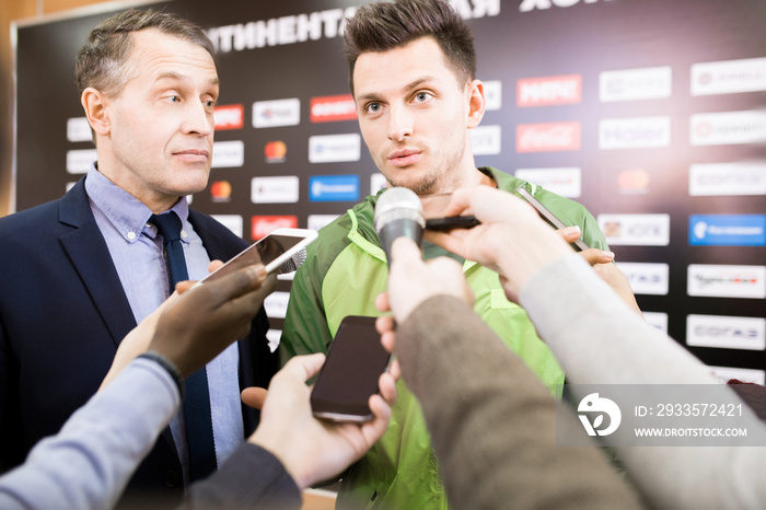 Portrait of handsome young sportsman and his manager giving interview to group of journalists during press conference