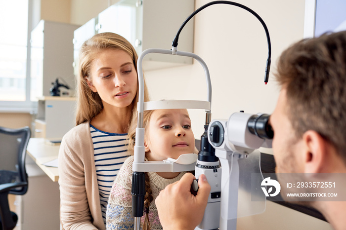 Portrait of cute little girl siting on mothers lap  looking at slit lamp machine during medical check up in eye clinic