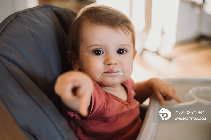 Baby girl with dirty lips pointing by finger directly to camera, sitting on high-chair. Baby care. Family care. Portrait of infant boy seated on highchair.