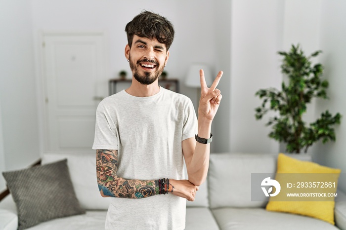 Hispanic man with beard at the living room at home smiling with happy face winking at the camera doing victory sign with fingers. number two.