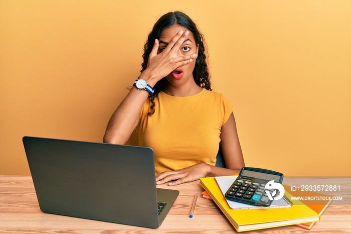 Young african american girl working at the office with laptop and calculator peeking in shock covering face and eyes with hand, looking through fingers with embarrassed expression.