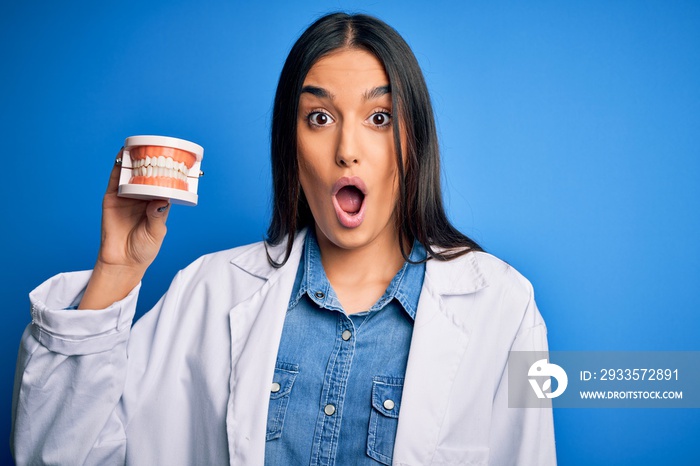 Young beautiful brunette dentist woman wearing coat holding plastic teeth prosthesis scared in shock with a surprise face, afraid and excited with fear expression