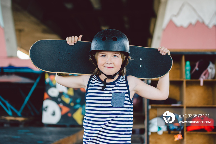 Portrait of handsome caucasian boy athlete skateboarder in protective helmet with skateboard in hands looking at camera on background of skate park. A child and an active hobby, sports and health