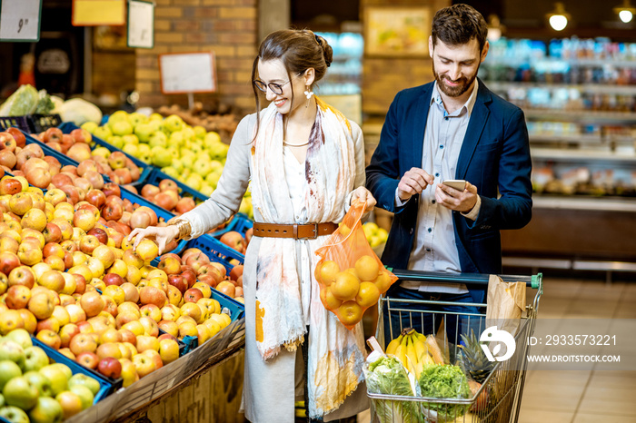 Young and happy couple buying fresh fruits and vegetables standing together with shopping cart in the supermarket