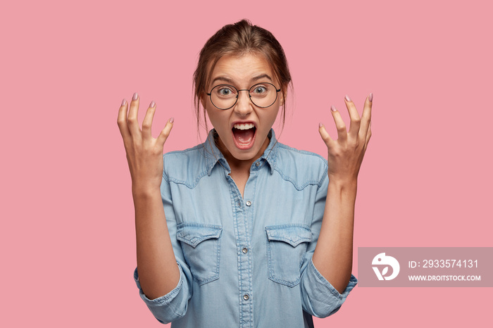 Photo of annoyed young woman gestures angrily, shouts with annoyance at friend, expresses negative feelings, dressed in stylish denim shirt, models indoor. Irritated teacher screams at noisy children