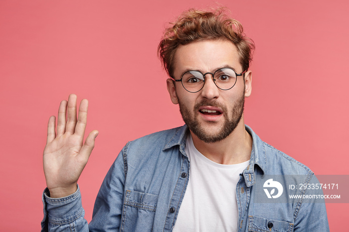 Indignant man wears jean shirt, waves with palm as tries to remind about himself. Active male student being puzzled, wants to give answer on question. Confused adult waves with hand in pink studio