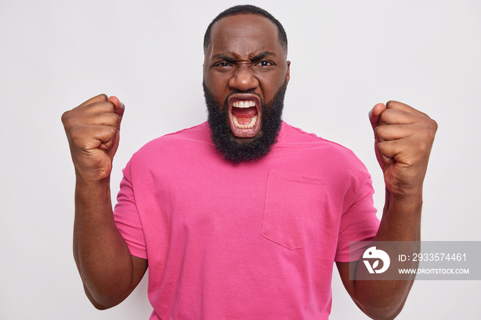 Anger concept. Outraged dark skinned bearded man clenches fists gestures furious screams from annoyance tired of problems expresses negative emotions wears pink t shirt isolated over white wall