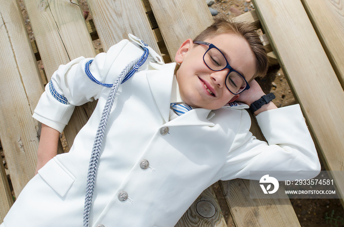 Happy child resting on a wooden plank dressed in communion suit. Lying down on wooden table wearing a white sailor suit