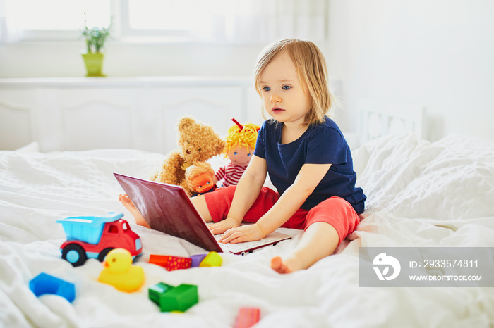 Toddler girl with laptop and toys in bed