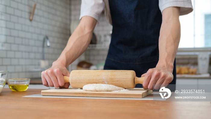 Unrecognizable young man using a rolling pin to roll over bread doughs on wooden table in kitchen.