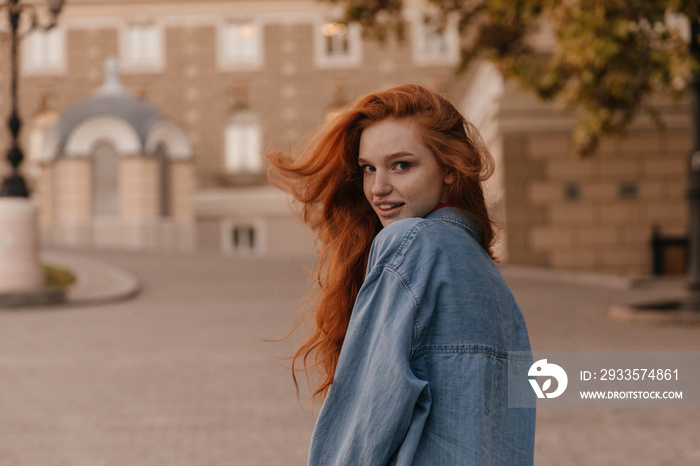 Portrait from back of young foxy lady posing outdoors. Gorgeous redshirted woman in denim shirt, looking into camera and smiling against city landmark background