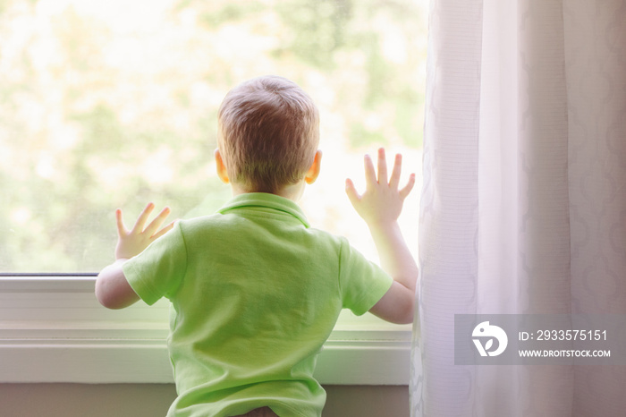 Portrait of white Caucasian child boy at home looking out of window outside. View from back. Kid waiting for someone