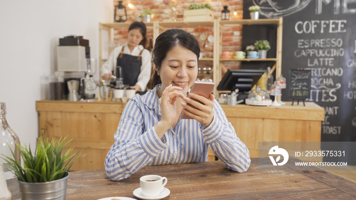 beautiful asian woman worker reading text message on mobile phone at coffee shop. happy girl customer smiling looking at smartphone screen with cup of tea. waitress in counter making client order.