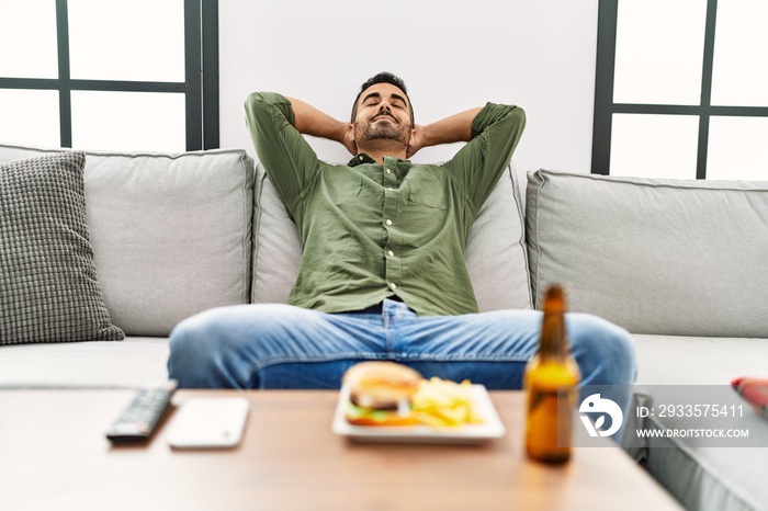 Young hispanic man relaxed with hands on head eating hamburger at home