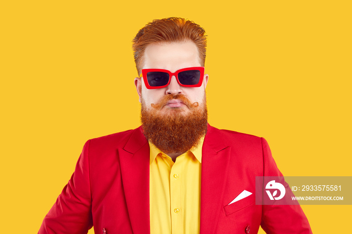 Funny stylish extravagant bearded chubby man pretending to be serious on vivid yellow background. Humorous red-haired guy in red jacket and sunglasses with funny serious and menacing look looks at you