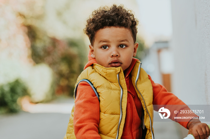 Outdoor portrait of cute little toddler boy playing in the park on a nice sunny day, wearing orange hoody jacket and yellow vest coat