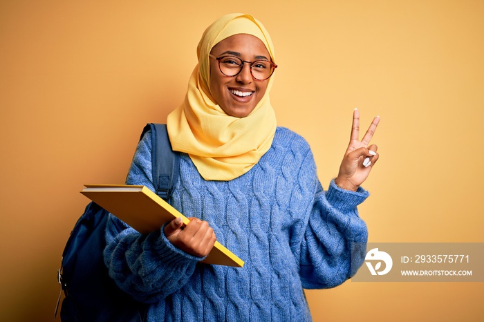 Young African American student woman wearing muslim hijab and backpack holding book smiling looking to the camera showing fingers doing victory sign. Number two.