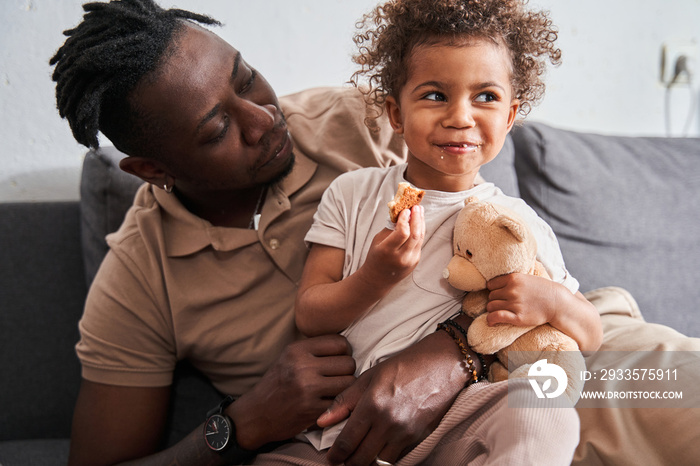 Man looking at his little curly daughter while sitting with her at the sofa